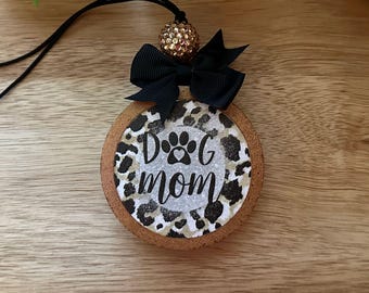 May include: A round cork ornament featuring a leopard print background with a central silver glitter circle. The text reads DOG mom with a paw print replacing the letter O. A black ribbon bow and a gold rhinestone bead sit at the top.