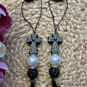 May include: Two phone charms featuring a black cross with a white leopard print pattern and a small black heart in the center. Each charm includes a large white pearl-like bead, a smaller black rhinestone-encrusted bead, and a black cord loop.