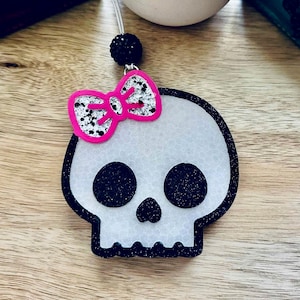 May include: A skull-shaped hanging ornament featuring a white textured face with black glittery eye sockets and a black glittery outline. A bright pink bow with black paint splatters is attached to the top left, and a black glittery bead sits above.