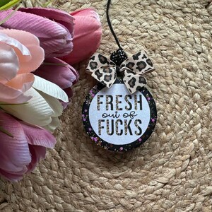 May include: A round hanging air freshener with a black border featuring pink and purple glitter accents. The center is white with the text FRESH out of FUCKS in black and leopard print lettering. A leopard print bow sits at the top with a black cord.