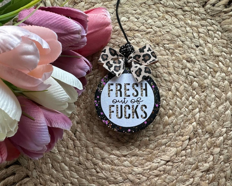 May include: A round hanging air freshener with a black border featuring pink and purple glitter accents. The center is white with the text FRESH out of FUCKS in black and leopard print lettering. A leopard print bow sits at the top with a black cord.