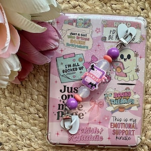 May include: A pink Kindle case with a clear cover, decorated with stickers and a beaded charm. The charm includes a miniature Kindle, purple beads, and a white flip-flop charm. Text on the case includes "Books are Therapy" and "Bookish Baddie."