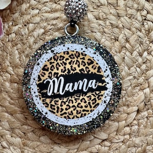 May include: A circular badge featuring a leopard print pattern with a black horizontal brushstroke across the center containing the white text Mama. The badge has a black border with multi-colored glitter and a silver-toned bead attachment.