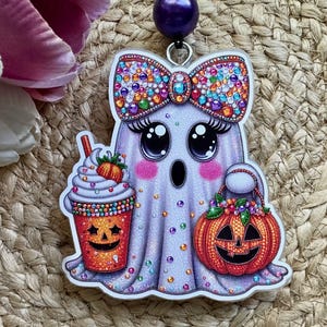 May include: A ghost-shaped pendant featuring a light purple body with colorful embedded gems, a large bow, and long eyelashes. The ghost holds an orange jack-o'-lantern bucket and a drink cup with a straw, both decorated with festive patterns.