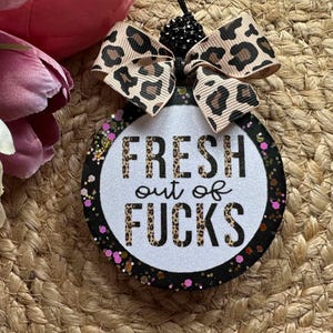 May include: A circular hanging ornament with a white glittery center featuring the black text FRESH out of FUCKS in a leopard print pattern. The border is black with pink and gold glitter accents, topped with a leopard print ribbon bow.