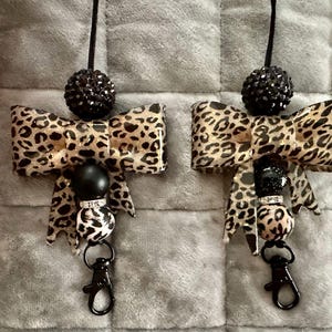 May include: Two matching lanyards featuring a leopard print bow, a large black glitter bead, a solid black bead, a silver-toned rhinestone spacer, and a smaller leopard print bead, all finished with a black metal lobster clasp and cord.
