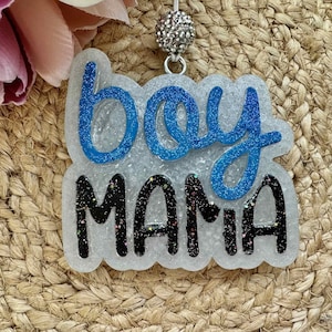 May include: A decorative resin badge featuring the word boy in blue glitter script above the word MAMA in black glitter block letters. The background is a translucent, glitter-flecked resin shape attached to a silver-toned metal ball bead charm.