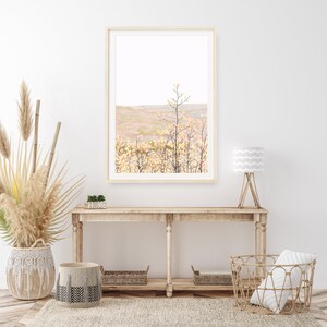 K&ouml;nnte beinhalten: Ein gerahmtes Foto eines Feldes mit hohem Gras und einer einzelnen, hohen Pflanze in der Mitte. Das Foto h&auml;ngt an einer wei&szlig;en Wand &uuml;ber einem Holztisch mit zwei K&ouml;rben und einer Lampe.