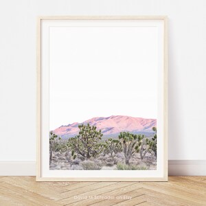 May include: A framed print depicting a desert scene with Joshua trees and a pink mountain range against a bright sky. The artwork is displayed on a wooden floor, leaning against a white wall. The print has a light wood frame.