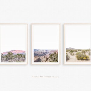 May include: Three framed landscape prints showcasing desert scenes. The prints depict a mountain range, a canyon, and a desert landscape with sparse vegetation. The frames are light-coloured, and the overall aesthetic is minimalist and serene.