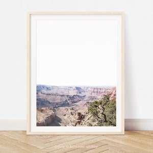 May include: A framed print depicting the Grand Canyon. The image shows layers of red and brown rock formations beneath a bright sky. A small tree is visible on the right. The print is in a light wood frame.