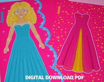 Op de afbeelding: Een printbare papieren poppenset met een meisje genaamd Annie. De pop heeft blond haar en draagt een turquoise jurk. De set bevat een roze jurk met een gouden accent.