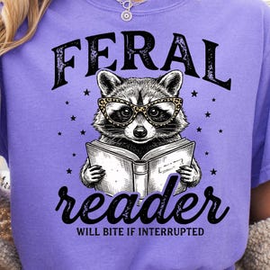 May include: A lilac-colored t-shirt with a graphic of a raccoon wearing glasses and reading a book. The text "FERAL reader WILL BITE IF INTERRUPTED" is above and below the raccoon illustration.