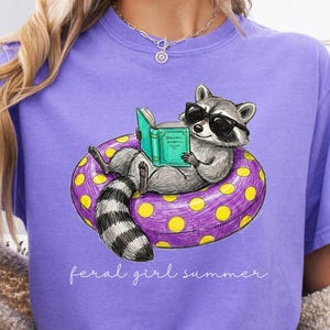 May include: A lilac-colored t-shirt featuring a cartoon raccoon wearing sunglasses and reading a book while floating on a purple and yellow inner tube. The text "feral girl summer" is written in cursive below the graphic.