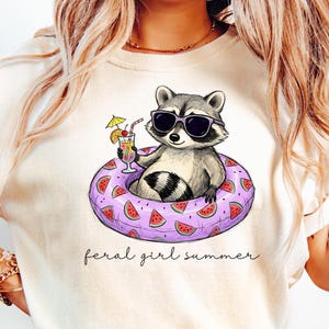 May include: Cream-colored t-shirt featuring a raccoon wearing sunglasses and floating in a watermelon-decorated inner tube. The raccoon is holding a cocktail. The text "feral girl summer" is written below the image.