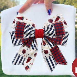 Op de afbeelding: Een grote, decoratieve strik met een patriottisch thema. De strik heeft een patchwork-ontwerp met rode, witte en blauwe patronen, waaronder strepen, ruiten en stermotieven. Een rode band vormt het midden. De strik wordt gepresenteerd op een wit oppervlak.