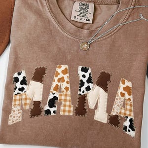 May include: A brown Comfort Colors t-shirt with the word "MAMA" appliqued across the front in a patchwork design. The letters feature various patterns, including cow print, gingham, and floral prints. A silver necklace with a small pendant rests on the shirt.