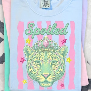 Op de afbeelding: Lichtblauw T-shirt met een afbeelding van een gekroonde luipaard, het woord "Spoiled", roze strepen en sterren. Het shirt is opgevouwen op andere pastelkleurige shirts.