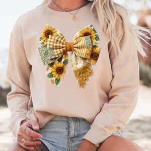 May include: A beige sweatshirt featuring a large bow design. The bow is composed of various patterned fabrics, including yellow gingham, floral, and dotted patterns. Sunflowers are incorporated into the bow's design, adding a touch of nature and color to the garment.