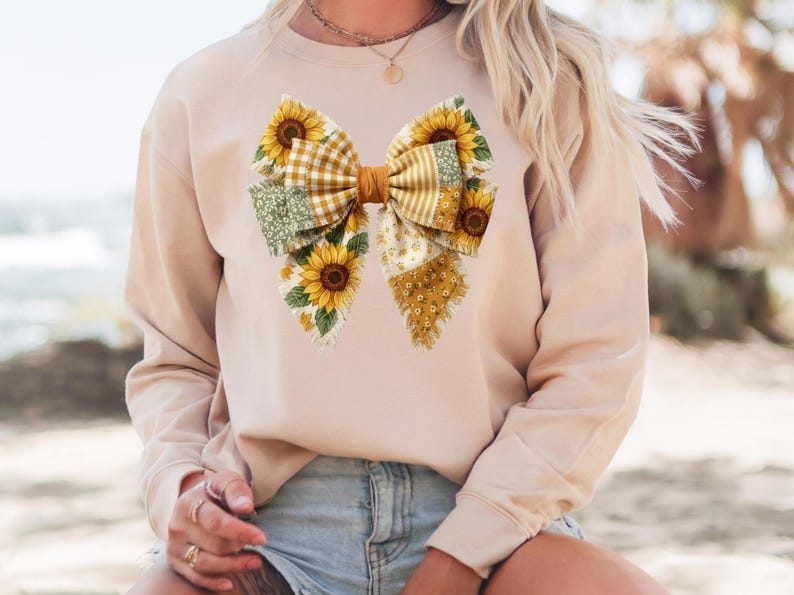 May include: A beige sweatshirt featuring a large bow design. The bow is composed of various patterned fabrics, including yellow gingham, floral, and dotted patterns. Sunflowers are incorporated into the bow's design, adding a touch of nature and color to the garment.