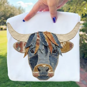 Puede incluir: Bolso tote blanco con un diseño de patchwork de la cara de una vaca de las Tierras Altas. La cara de la vaca está hecha de mezclilla y otras telas, con cuernos color beige y una franja desgreñada y multicolor. El bolso tote es sostenido por una mano.