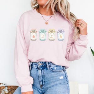 May include: A light pink sweatshirt featuring a watercolor design of four jars with Easter-themed illustrations. The jars are decorated with ribbons and contain an egg, bunny, flowers, and a carrot. The model is wearing blue jeans.