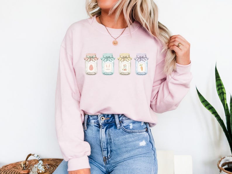May include: A light pink sweatshirt featuring a watercolor design of four jars with Easter-themed illustrations. The jars are decorated with ribbons and contain an egg, bunny, flowers, and a carrot. The model is wearing blue jeans.