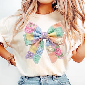 Pu&ograve; includere: T-shirt color crema con un grande design a fiocco colorato. Il fiocco &egrave; realizzato in tessuto patchwork dai colori pastello, tra cui rosa, blu, giallo e verde, con motivi floreali e a quadretti. Il fiocco &egrave; il punto focale della maglietta.