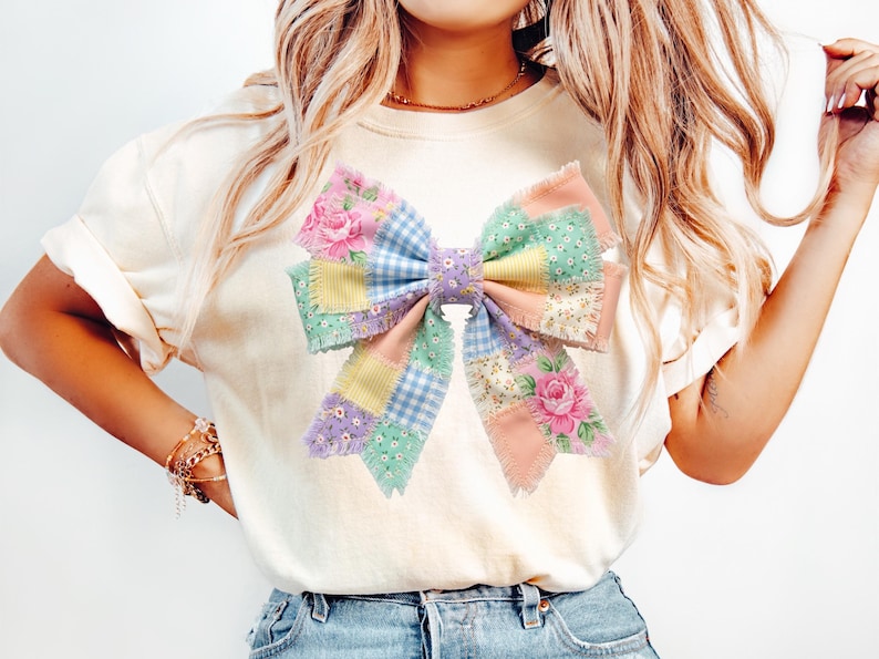 Pu&ograve; includere: T-shirt color crema con un grande design a fiocco colorato. Il fiocco &egrave; realizzato in tessuto patchwork dai colori pastello, tra cui rosa, blu, giallo e verde, con motivi floreali e a quadretti. Il fiocco &egrave; il punto focale della maglietta.