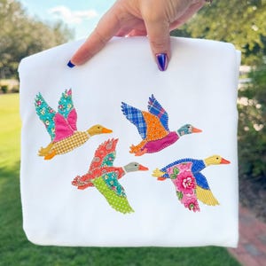 May include: White sweatshirt featuring four colorful embroidered ducks in flight. Each duck has wings made of patterned fabric in shades of pink, blue, orange, and yellow. The ducks have orange beaks.