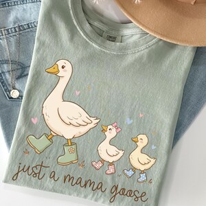 May include: Sage green t-shirt featuring a cartoon goose and two goslings, all wearing boots. The text "just a mama goose" is below the illustration. The shirt is laid on top of blue jeans.