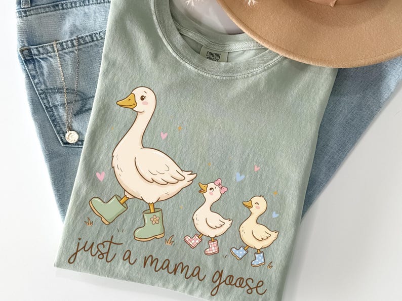 May include: Sage green t-shirt featuring a cartoon goose and two goslings, all wearing boots. The text "just a mama goose" is below the illustration. The shirt is laid on top of blue jeans.