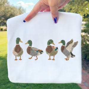 Pode incluir: Camisola branca com um bordado de quatro patos. Os patos têm cabeças verdes, corpos castanhos e asas em patchwork. A camisola é segurada por uma mão com verniz azul.