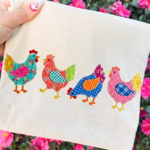 May include: Cream-colored fabric featuring four colorful, appliqued chickens. Each chicken has a unique pattern, including floral, paisley, and polka dots. The chickens are in shades of blue, pink, red, and yellow, with orange feet and red combs.