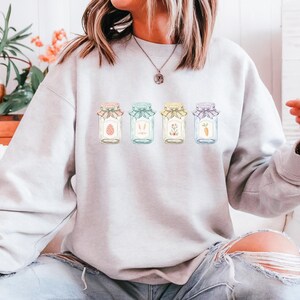 May include: A light gray sweatshirt featuring four jars with Easter-themed designs. Each jar has a different pastel-colored lid and contains an Easter egg, bunny ears, flowers, and a carrot. The sweatshirt is worn by a person.