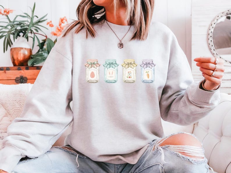 May include: A light gray sweatshirt featuring four jars with Easter-themed designs. Each jar has a different pastel-colored lid and contains an Easter egg, bunny ears, flowers, and a carrot. The sweatshirt is worn by a person.