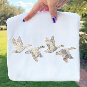 以下が含まれることがあります： 白いスウェットシャツに、飛んでいる4羽のガチョウの刺繍が施されています。ガチョウは、ベージュとグレーの色合いの模様のある翼と体を持っています。スウェットシャツは、青いマニキュアを塗った手で持たれており、緑の芝生の背景があります。