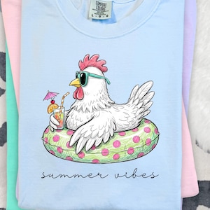 May include: Pale blue Comfort Colors t-shirt with a cartoon chicken wearing sunglasses and floating on a green and pink polka dot inflatable ring. The chicken holds a cocktail. The words "summer vibes" are written in script.