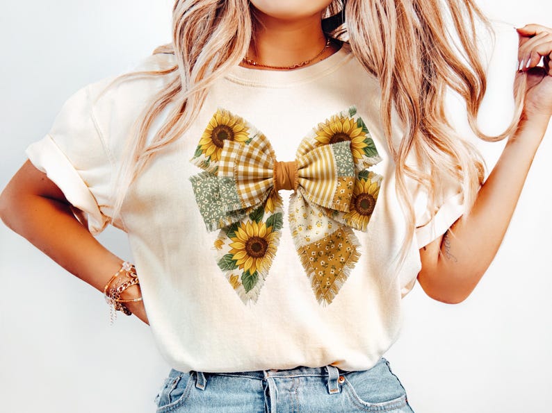 May include: Cream-colored t-shirt featuring a large bow design. The bow is made of various fabric patterns, including sunflowers, gingham, and floral prints. The bow has a brown center and is the focal point of the shirt.