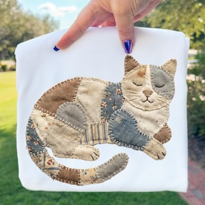 May include: A white pillow cover featuring a patchwork cat design. The cat is composed of various fabric scraps in shades of brown, blue, and cream, stitched together to create a whimsical, handcrafted aesthetic. The cat is lying down with its eyes closed.