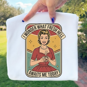 May include: White sweatshirt featuring a retro-style graphic of a woman holding a coffee cup, with the text "I WONDER WHAT FRESH HELL AWAITS ME TODAY". The graphic is in shades of red, yellow, and green.