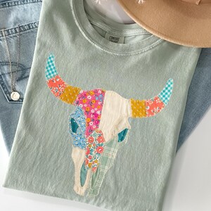 May include: A light green t-shirt featuring a colorful patchwork bull skull design. The horns are adorned with floral and gingham patterns. The shirt is paired with light blue jeans and a tan hat.