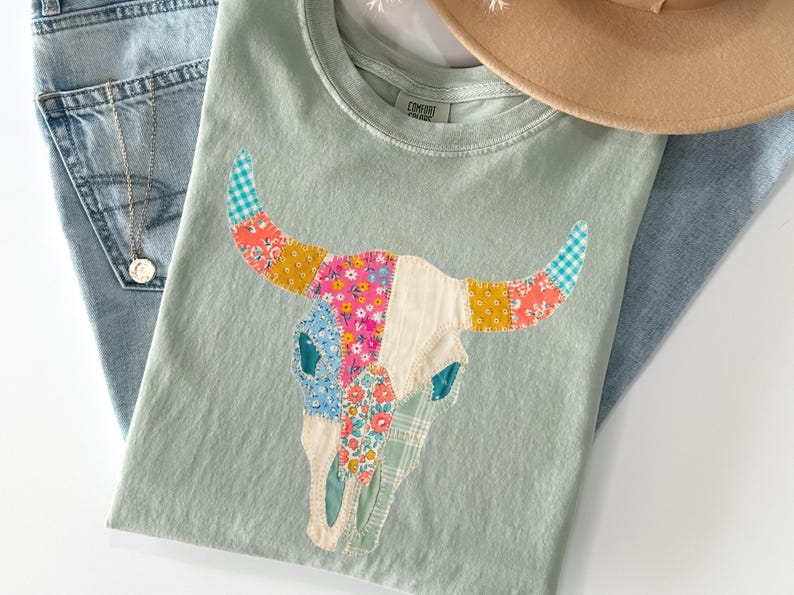 May include: A light green t-shirt featuring a colorful patchwork bull skull design. The horns are adorned with floral and gingham patterns. The shirt is paired with light blue jeans and a tan hat.