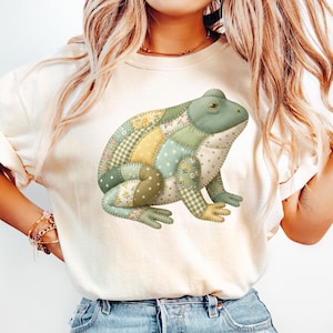 Könnte beinhalten: Cremefarbenes T-Shirt mit einem skurrilen Froschdesign. Der Frosch besteht aus verschiedenen Stoffmustern in Grün-, Gelb- und Weißtönen, wodurch ein Patchwork-Effekt entsteht. Der Frosch ist das zentrale Element des Shirts.