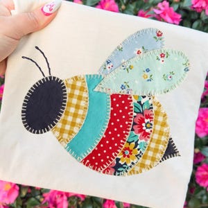 May include: A cream-colored fabric square featuring a colorful, appliqued bee design. The bee has a black head, yellow and white checkered body segments, turquoise, red polka dot, and floral patterned sections, and light blue floral wings. Black antennae complete the design.
