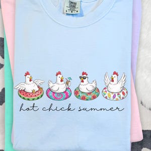 May include: Pale blue t-shirt featuring a graphic of four chickens on inflatable rings. The text "hot chick summer" is below the graphic. The shirt is folded. The chickens are on different designs of inflatable rings.