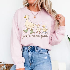 May include: A light pink sweatshirt featuring a cartoon illustration of a mama goose and two goslings, all wearing boots. The text "just a mama goose" is written below the illustration. The sweatshirt is paired with blue jeans.