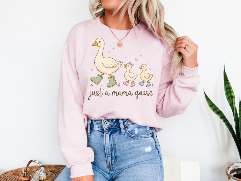 May include: A light pink sweatshirt featuring a cartoon illustration of a mama goose and two goslings, all wearing boots. The text "just a mama goose" is written below the illustration. The sweatshirt is paired with blue jeans.