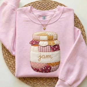 May include: A light pink sweatshirt featuring a patchwork jam jar design. The jar has a beige label with the word "jam" embroidered in a cursive font. A gold heart pendant hangs from a delicate necklace.