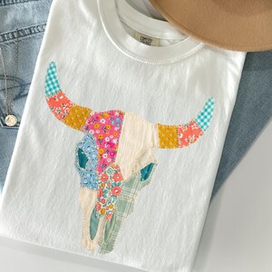 May include: White t-shirt featuring a colorful patchwork bull skull design. The skull is made of various patterned fabrics in shades of pink, blue, yellow, and green. The shirt is paired with blue jeans and a tan hat.